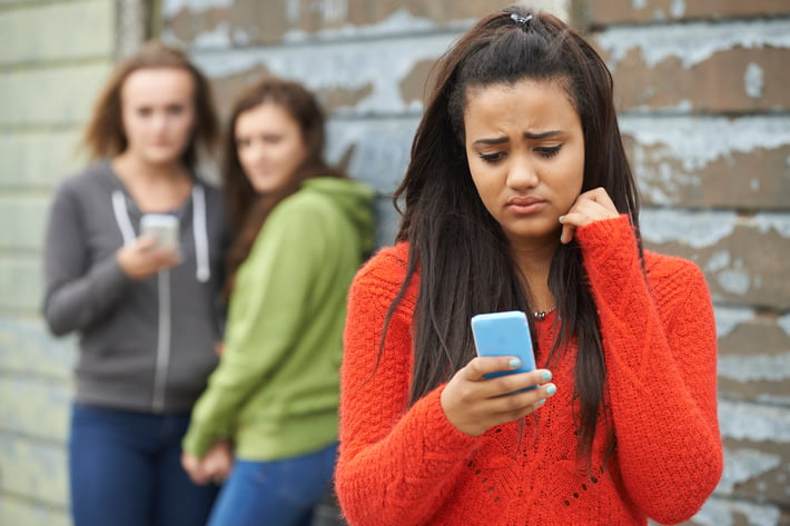 2 girls looking at other girl as she is upset reading something on her phone