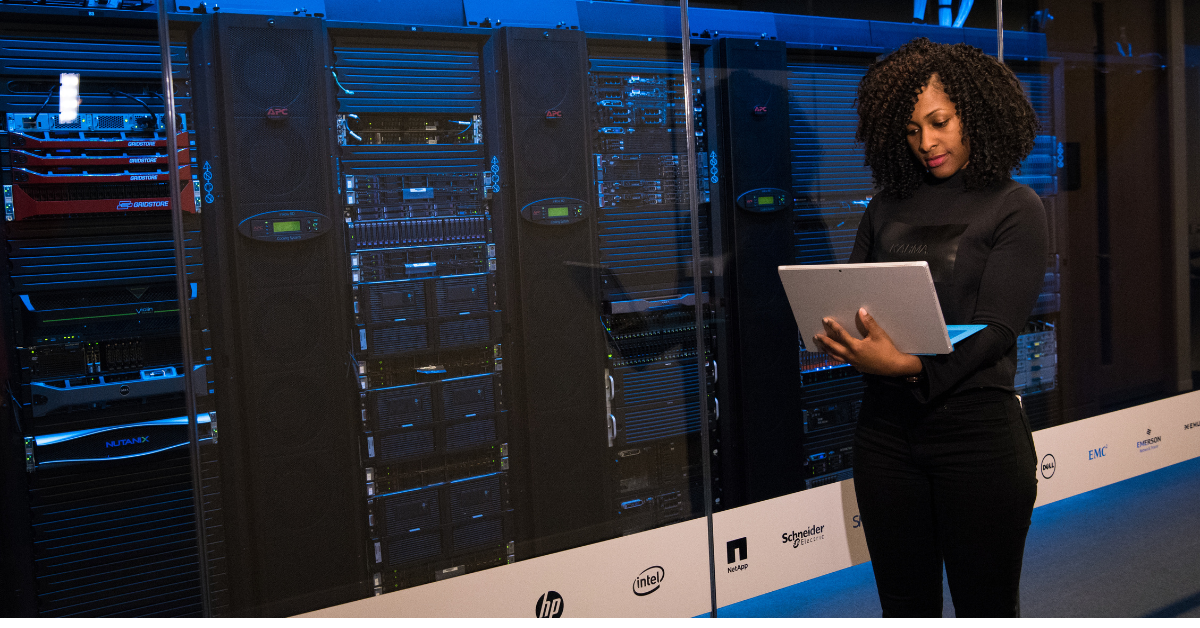 A woman with curly hair stands in a data center, viewing a laptop or tablet she's holding. She's dressed in a black turtleneck and pants, positioned in front of illuminated server racks 