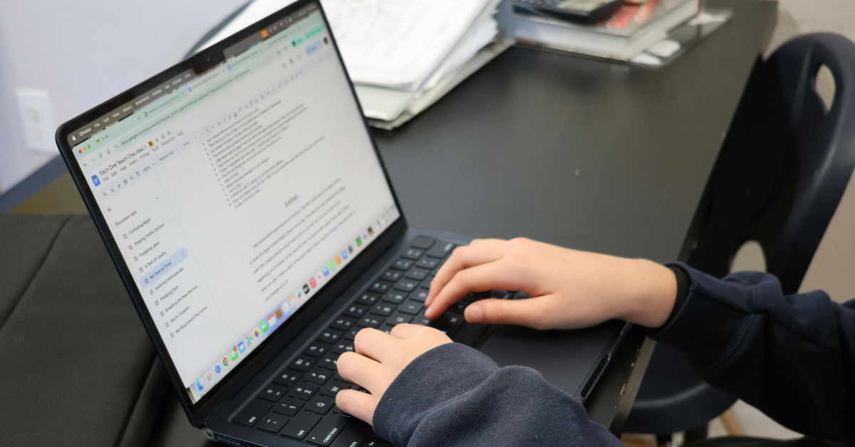 Student's hands typing on a laptop, working on an assignment in Google Docs at a classroom desk.