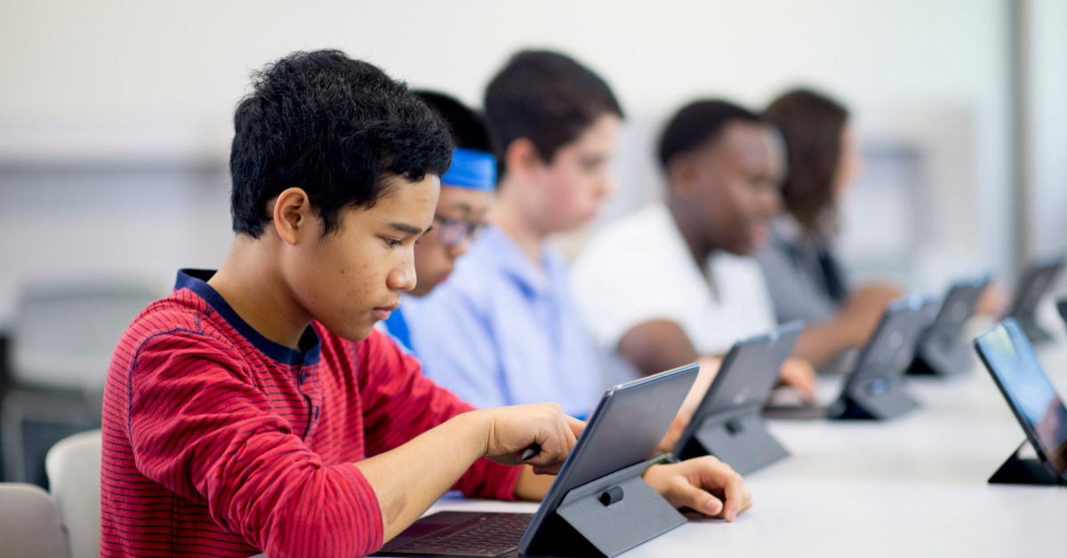 Students, seated in a row at desks, use tablets during class; in the foreground, a teenage boy in a red striped shirt taps his tablet screen.