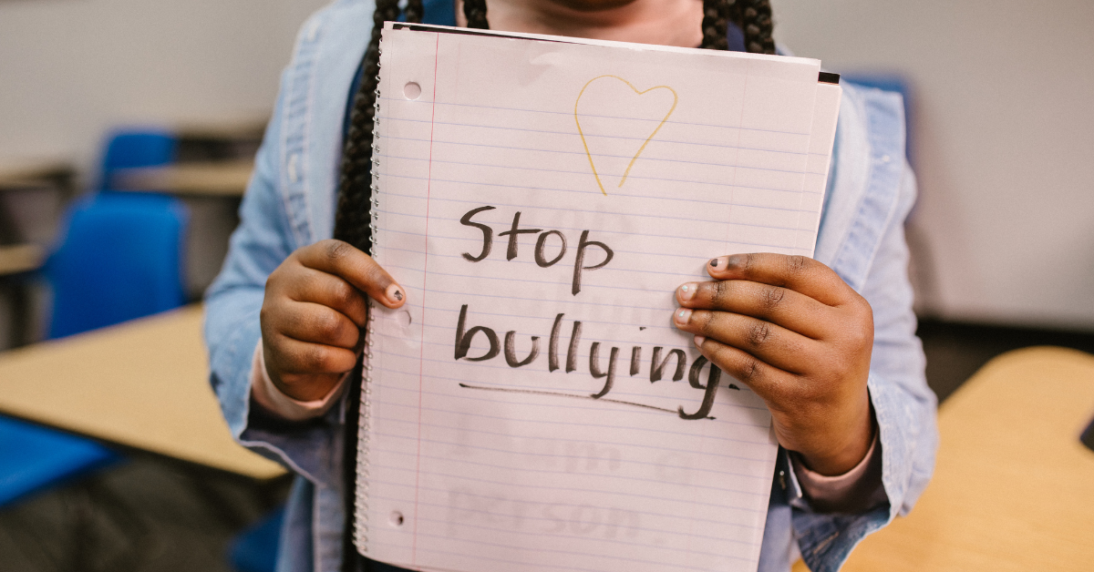 Student in a classroom holding up a handwritten notebook page reading 'Stop bullying' with a heart drawn above it