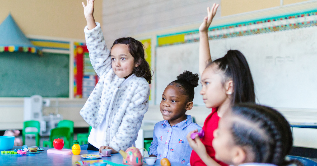 Young students raising their hands at a classroom activity table, with colorful educational materials and a chalkboard in the background.
