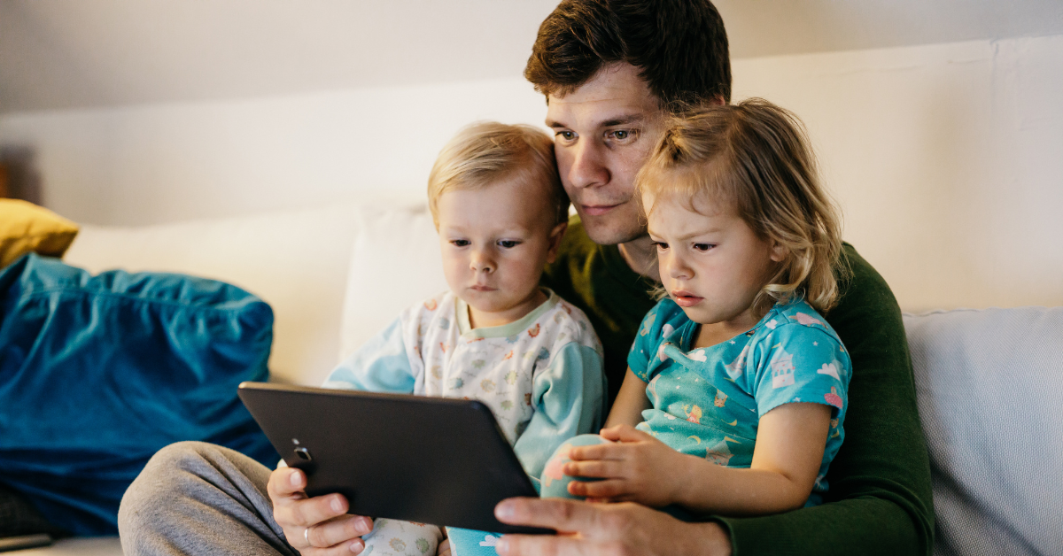 Father sitting with two toddlers on his lap, looking at a tablet together.