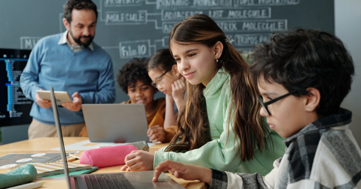 Middle school students use laptops in a classroom while a teacher monitors their digital activity on a tablet