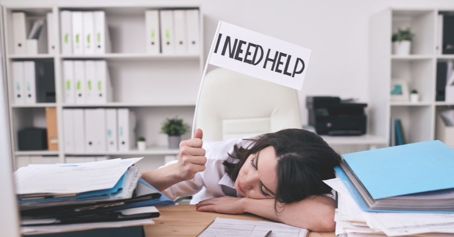 Young student resting her head on a messy desk amid papers, holding up a sign that says I Need Help.